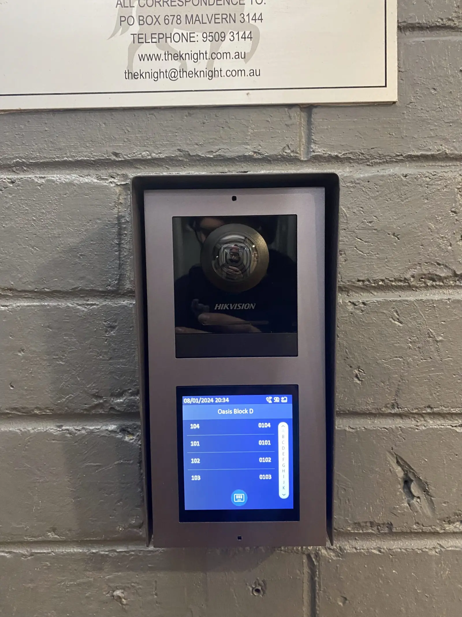 Video intercom panel with a directory screen listing unit numbers, mounted on a brick wall at a block entrance