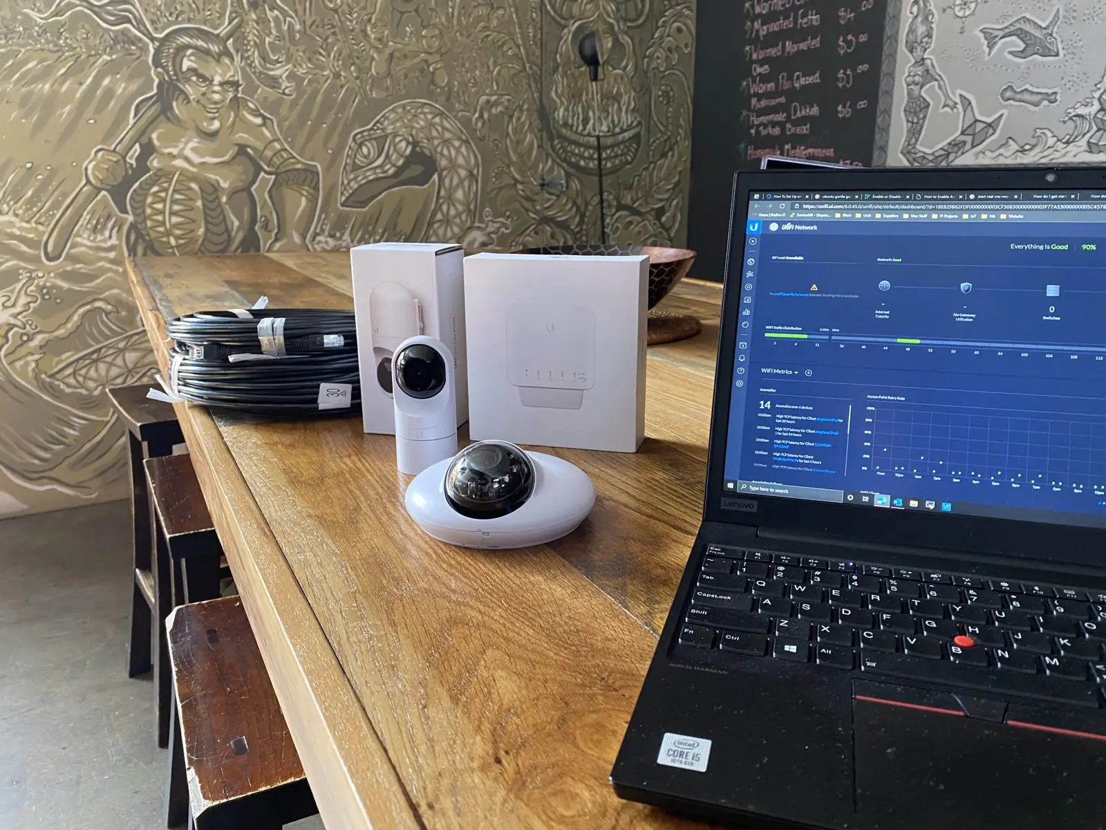 A UniFi Protect camera and its packaging on a venue table beside a laptop showing the controller, ready to commission